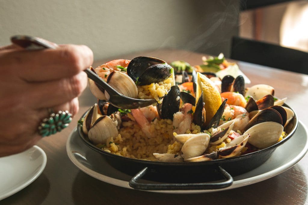 A person with light-medium skin tone, wearing a turquoise ring, uses a dark-colored spoon to lift a portion of a seafood paella from a black, shallow, round pan. The paella consists of yellow rice, mussels, clams, shrimp, and a lemon wedge. Steam rises from the dish. The pan sits on a white plate, which is on a wooden table.  A partially visible white plate is in the lower left corner. The background is blurred but shows a portion of a wooden table and a dark-colored chair.  The lighting is warm and highlights the food.