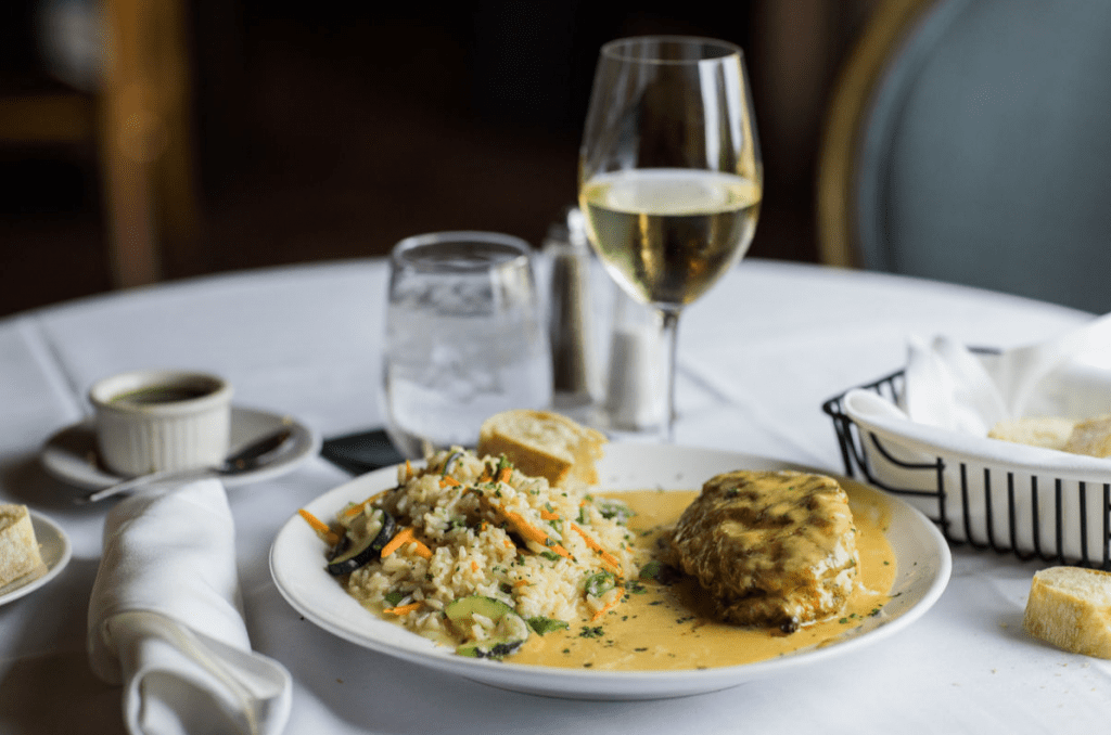 A white tablecloth covers a round table set for a meal. A white plate holds a portion of rice pilaf with mixed vegetables (zucchini, carrots, and possibly green beans) in a creamy sauce. Next to the rice is a piece of breaded and pan-fried chicken breast, also covered in the same creamy sauce. Garnished with fresh herbs. A glass of white wine sits to the right of the plate. To the left, there is a small white ramekin containing a dark green sauce, a folded white napkin, and a piece of bread on a small side plate. A glass of ice water is partially visible behind the plate. More bread is visible in a wire bread basket in the background. The background is blurred but shows a dark interior with hints of wood tones. The lighting is soft and warm, focusing on the food.